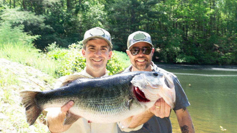 Taylor LeBlanc and Steve holding a big largemouth bass at the edge of a lake