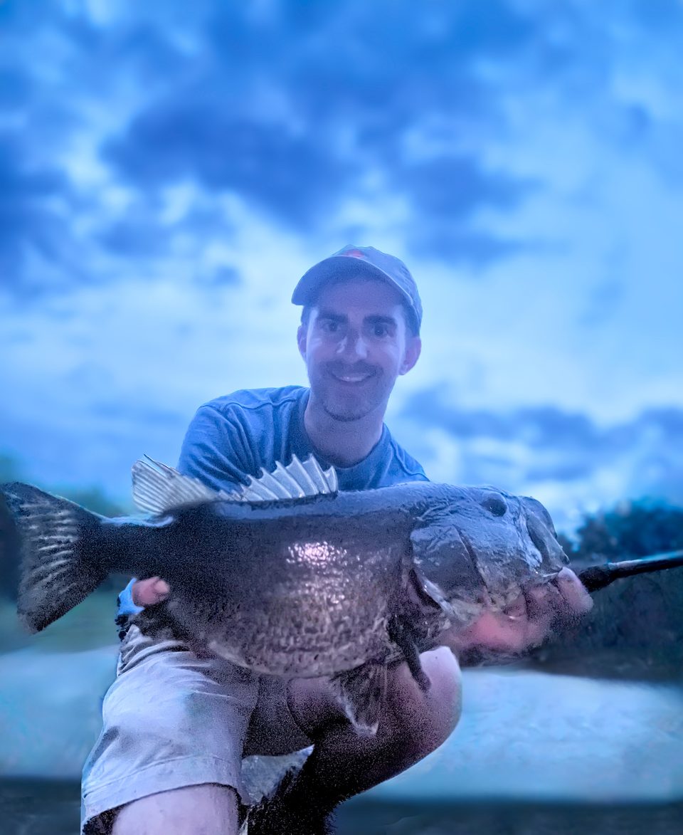 Taylor LeBlanc crouched at the water's edge with a largemouth bass at dusk