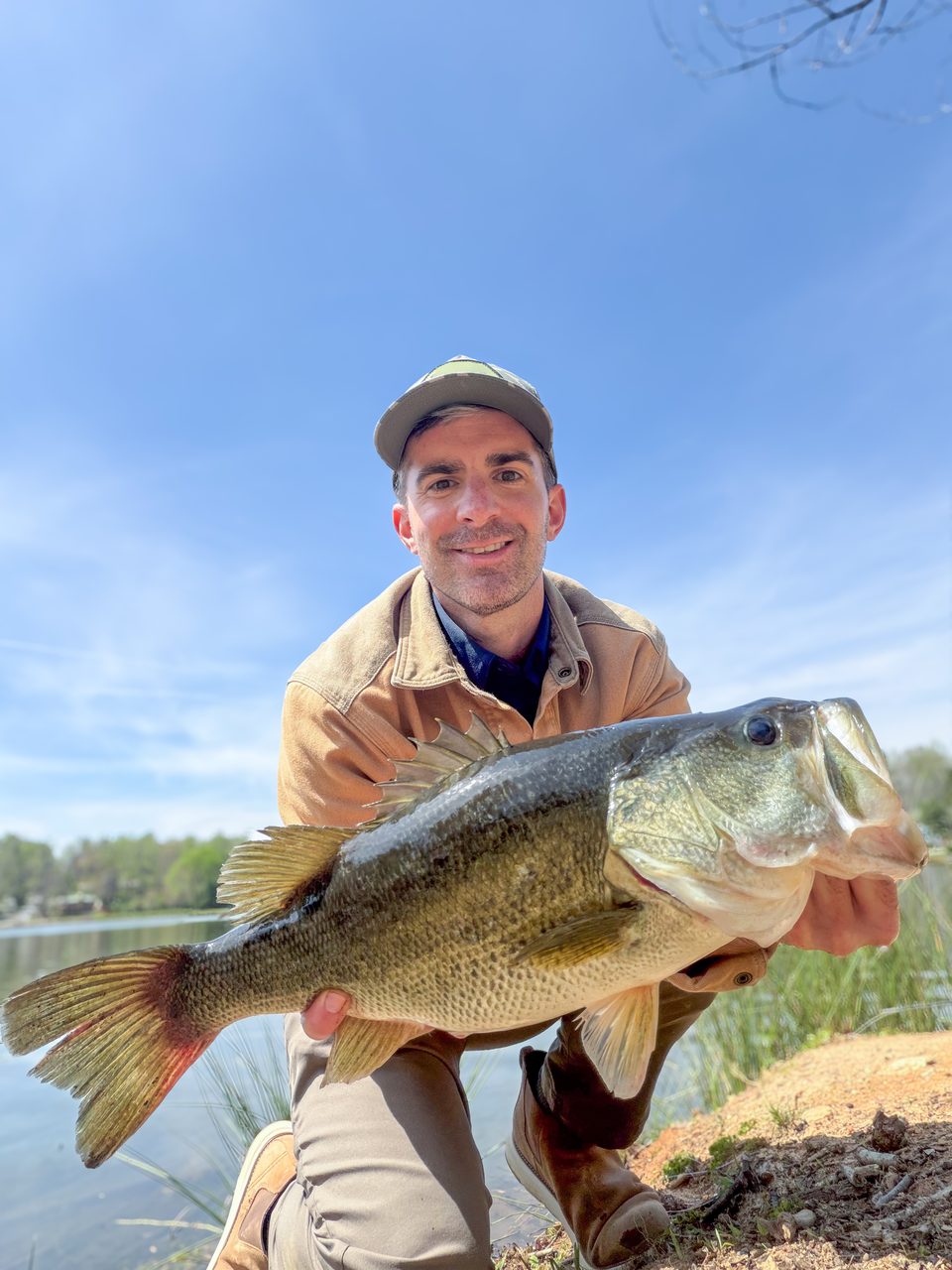 Taylor LeBlanc kneeling on a sandy bank with a largemouth bass under a blue sky