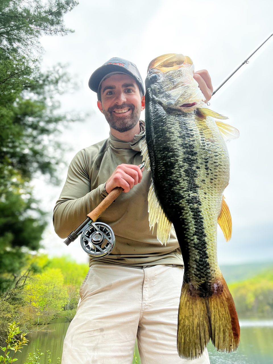 Taylor LeBlanc holding a largemouth bass up beside a fly rod